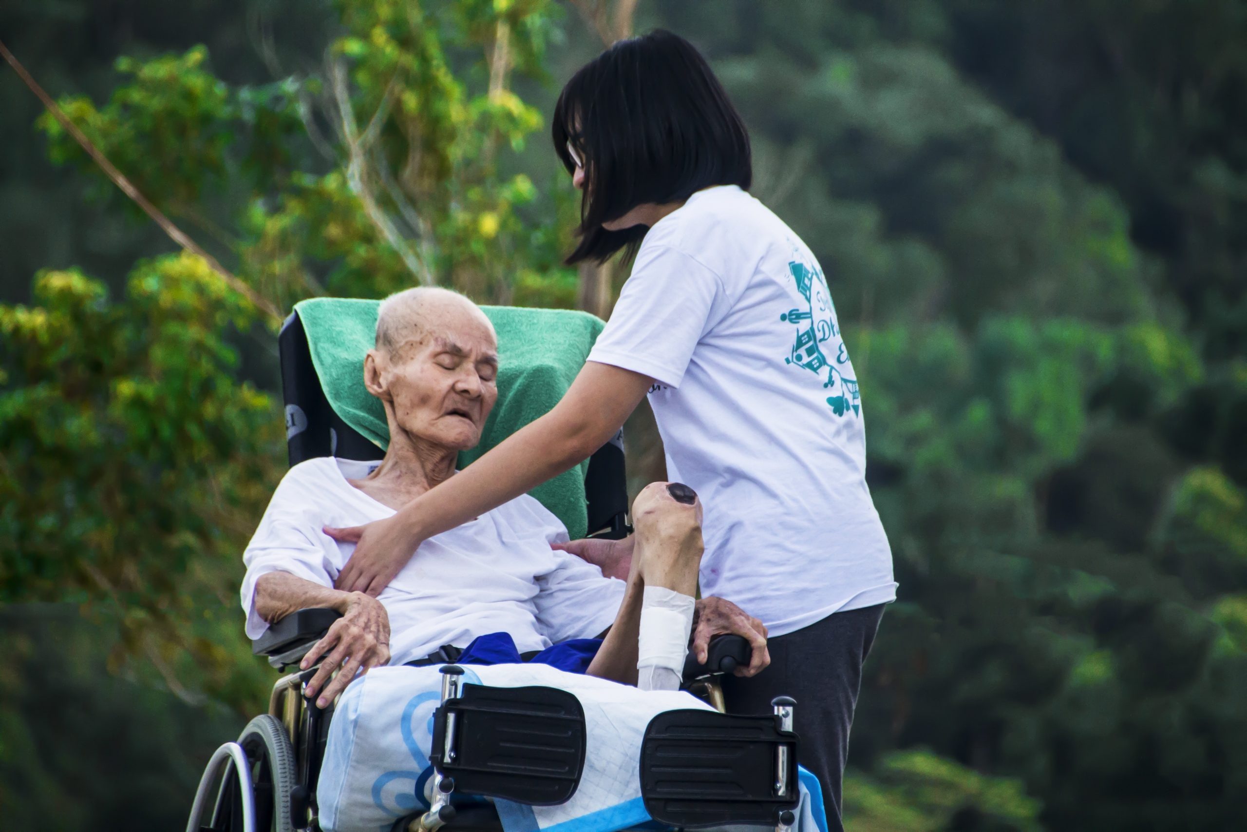 An elderly frail gentleman on a wheelchair with physical limitations is being taken care of by someone who could be a family member or a paid caregiver. You cannot tell...